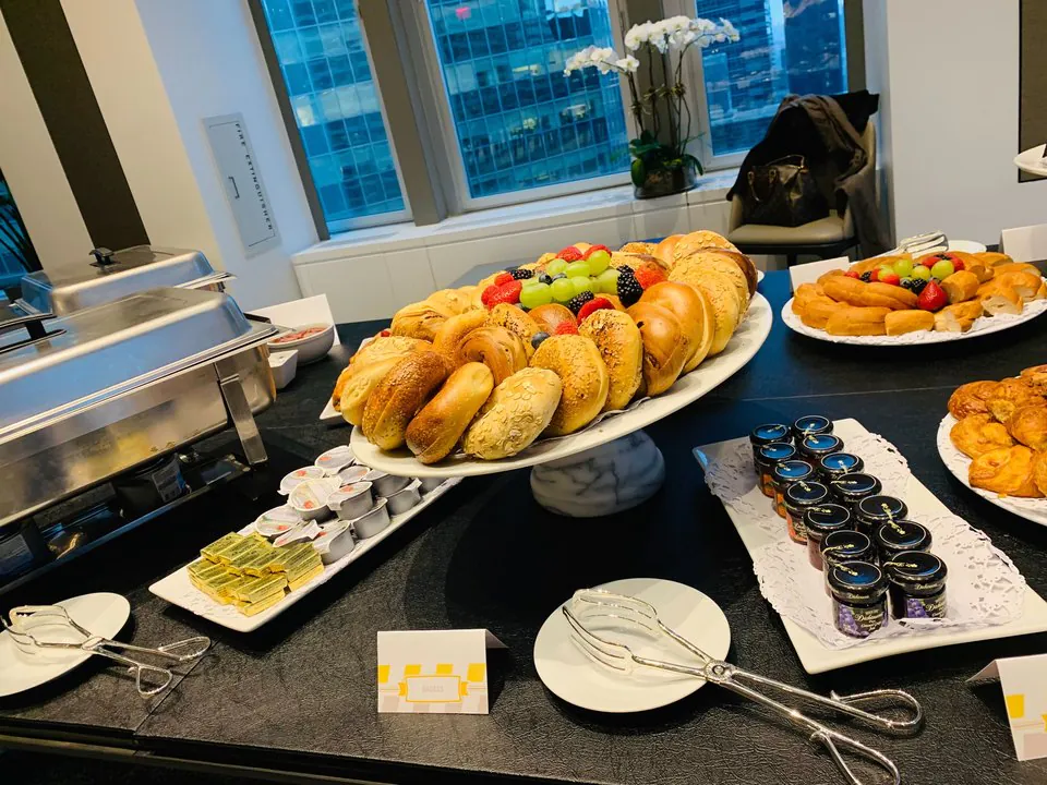 Assorted bagels arranged in a ring topped with fresh fruit on a marble pedestal, with butter, jam jars, and a chafing dish on an office buffet