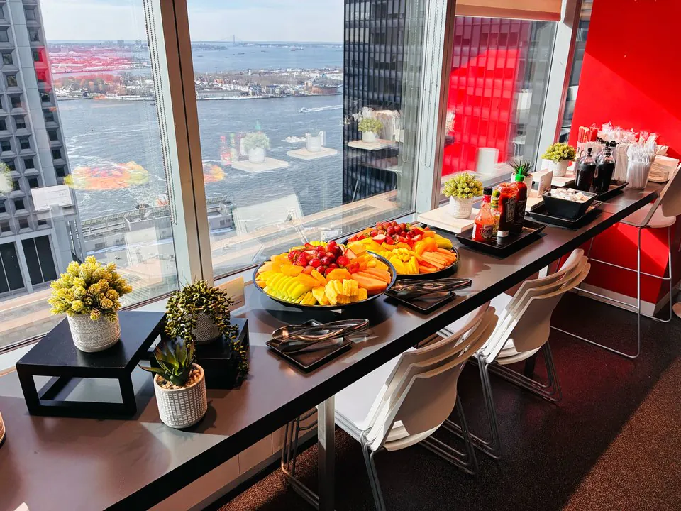 Bright high-rise office breakfast counter with platters of fresh fruit, beverages, and a red accent wall overlooking the water