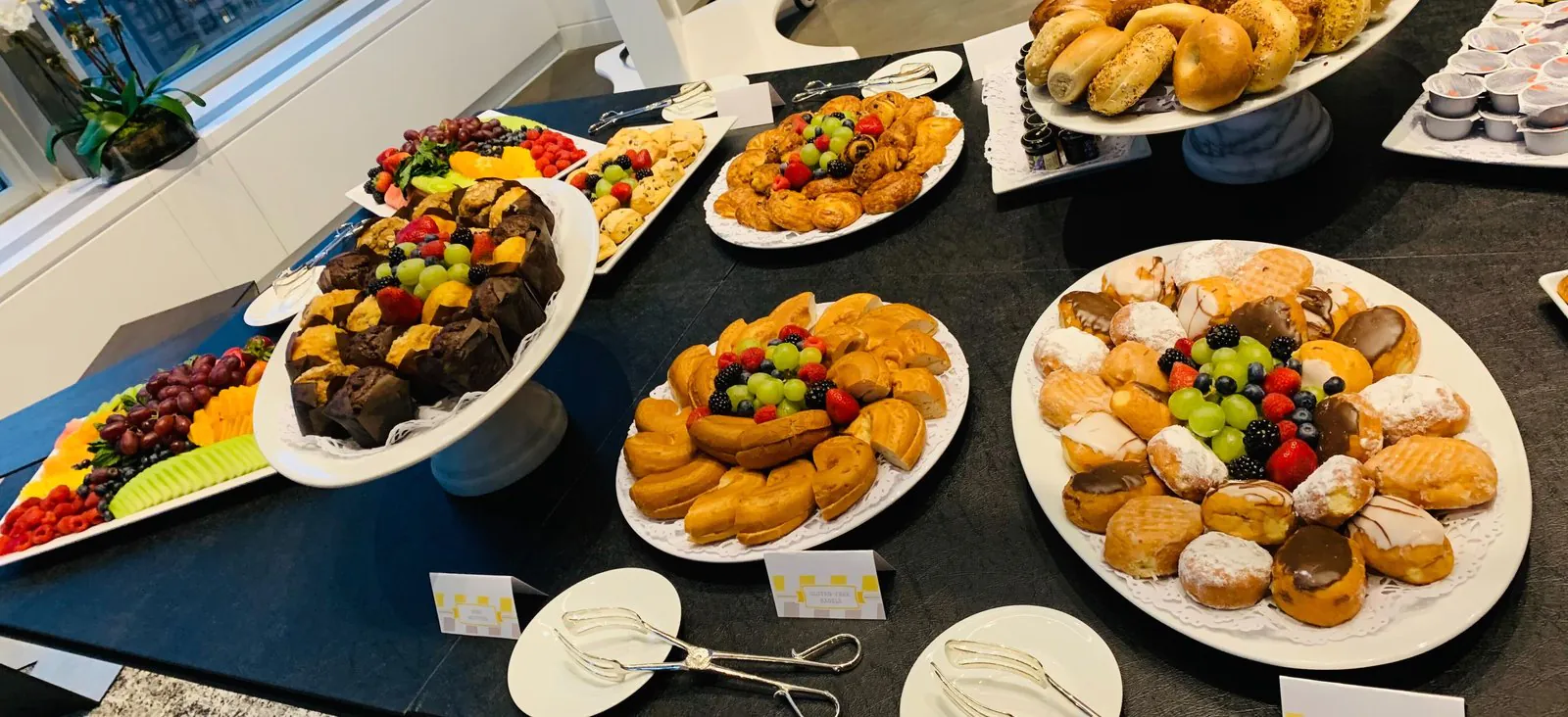 Lavish office breakfast spread of bagels, pastries, muffins, and fresh fruit on white pedestal platters in a high-rise office
