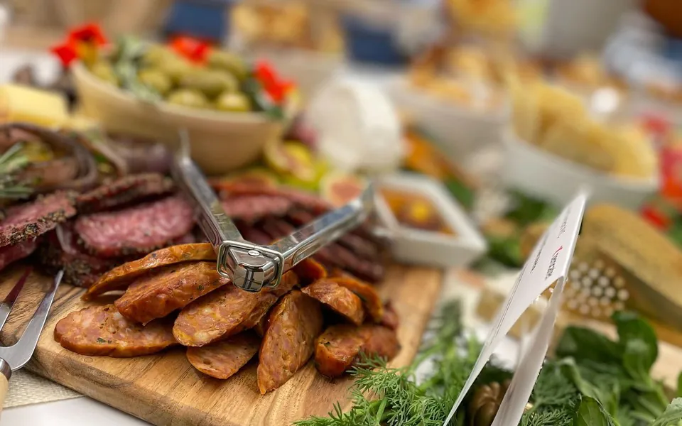 Close-up of a charcuterie board with sliced cured sausage and salami, olives, mozzarella, and fresh herbs on a wooden board at an office grazing table