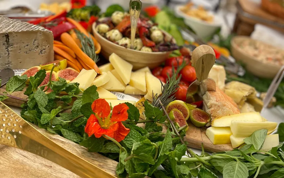 Office grazing board with sliced cheeses, carrots, cherry tomatoes, fresh figs, marinated mozzarella, mint, and edible flowers arranged on a wooden board