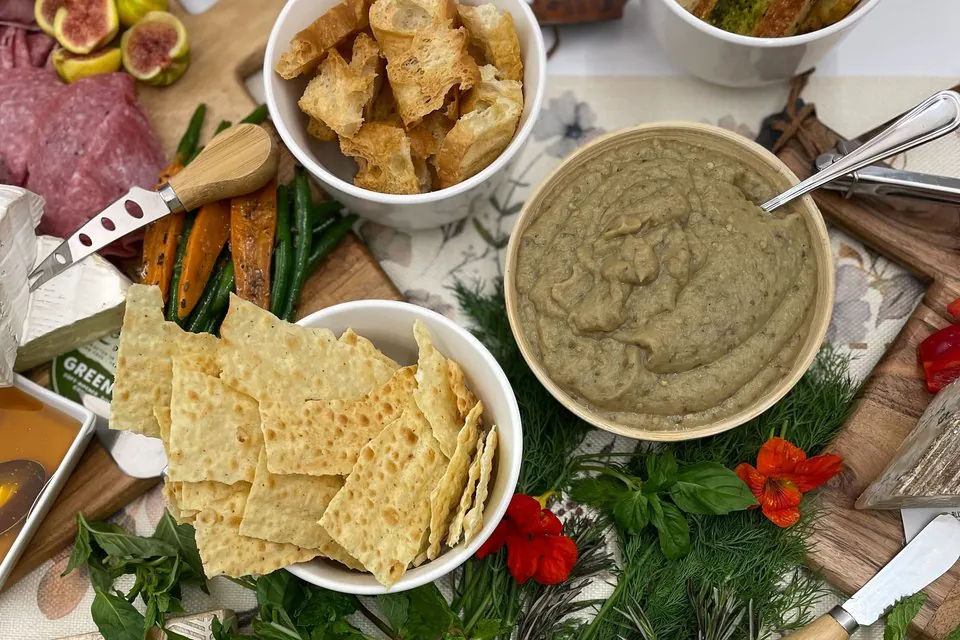 Overhead view of a happy hour dip spread with baba ganoush, crackers, crostini, crudite, fresh herbs, and charcuterie on a marble surface