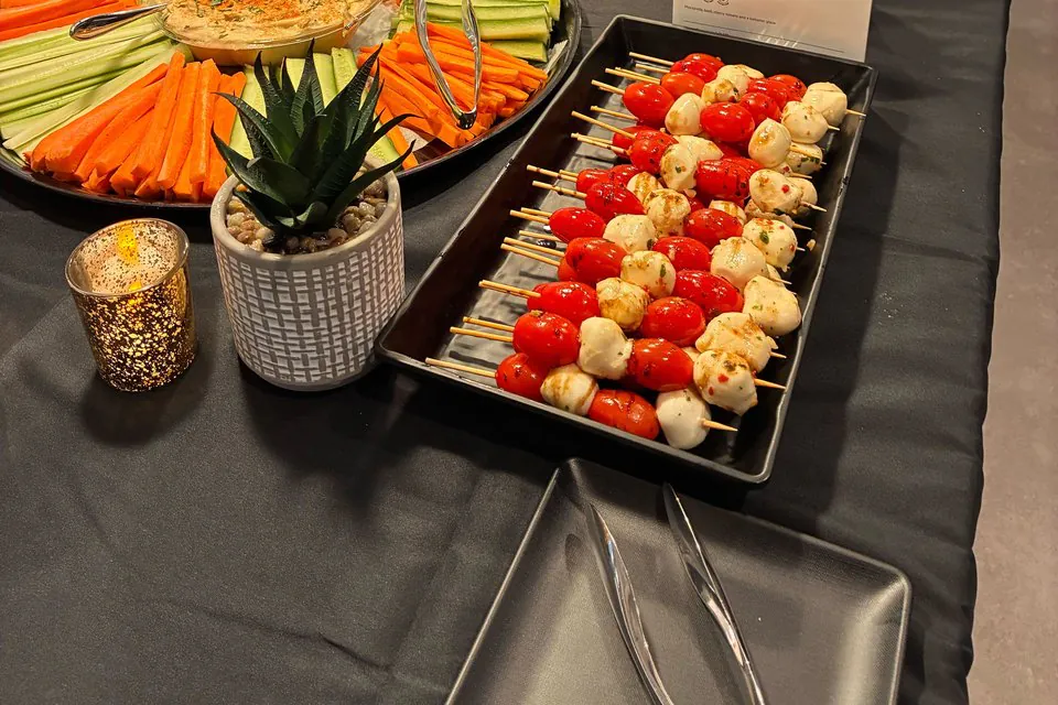 Office happy hour appetizer spread with caprese skewers of cherry tomato and mozzarella on a black tray beside a crudite platter of carrots and cucumber with hummus