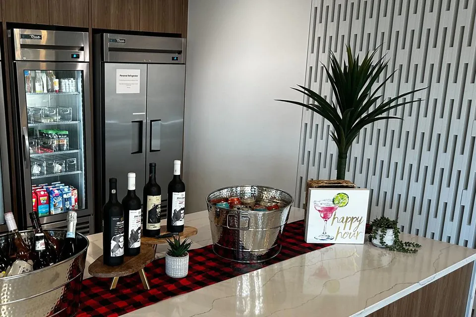 Self-serve office happy hour bar with hammered metal ice tubs of wine and canned beverages, wine bottles on a wood riser, and a small happy hour sign on a kitchen counter