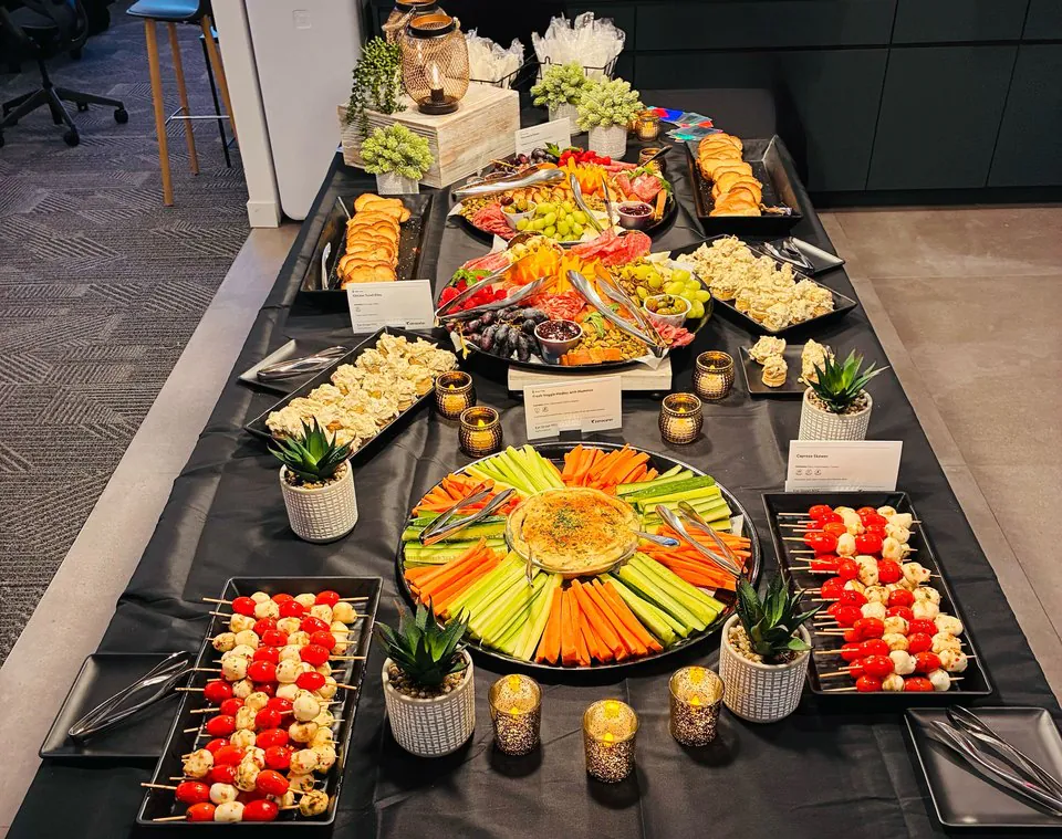 Symmetrical office happy hour spread with caprese skewers, a crudite sunburst with hummus dip, charcuterie, cheese, and fruit on a dark linen table