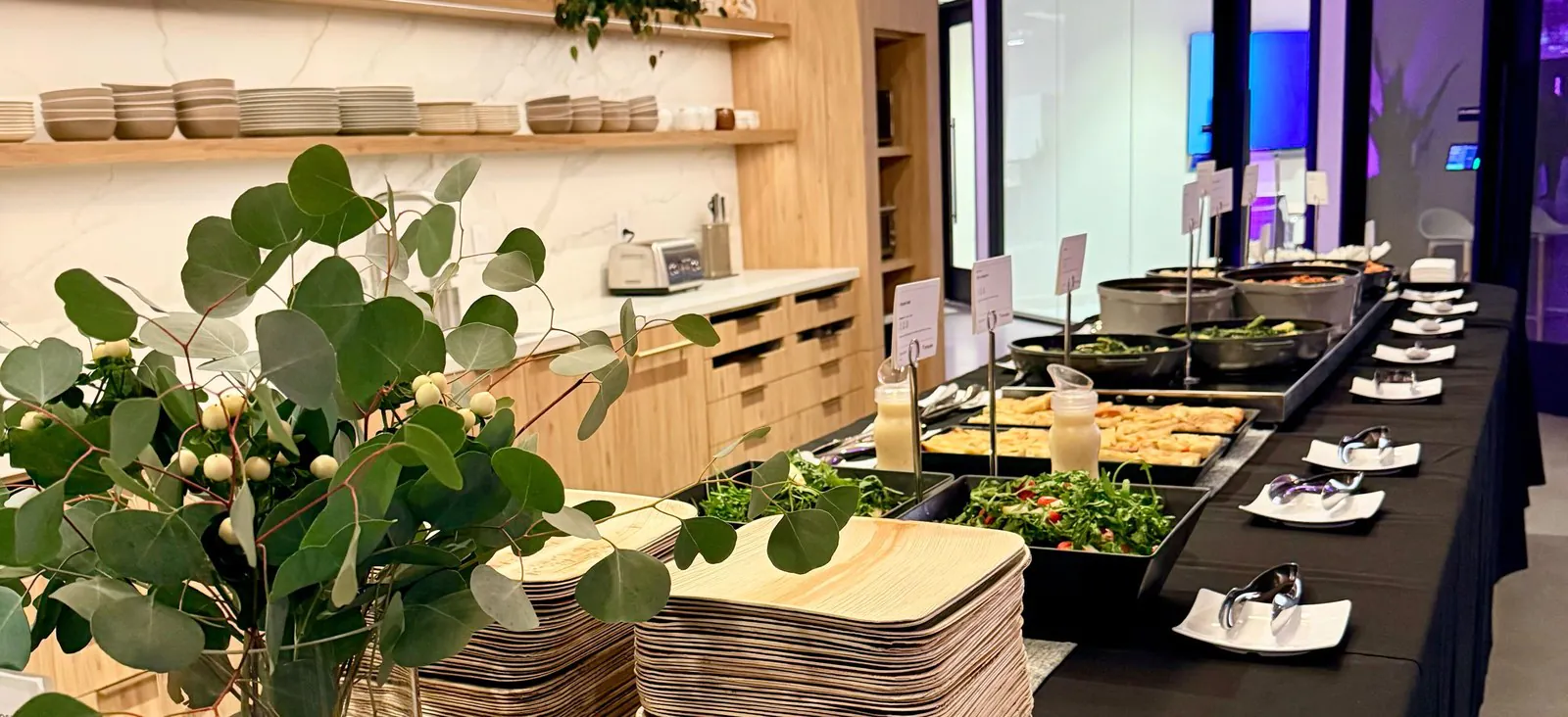 Healthcare staff appreciation catering buffet with stacked wood-look plates, a fresh herb centerpiece, and chafing trays of salads and pastries along a long office break-room table