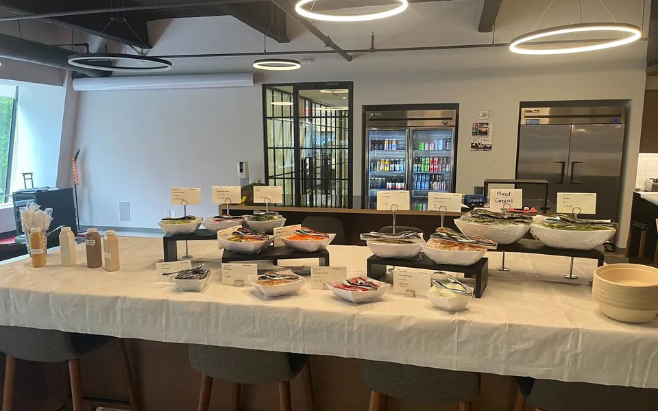 Build-your-own salad and grain bar in a modern office with tiered white bowls of mixed greens and toppings, dressings on the side, and a handwritten Mixed Greens sign