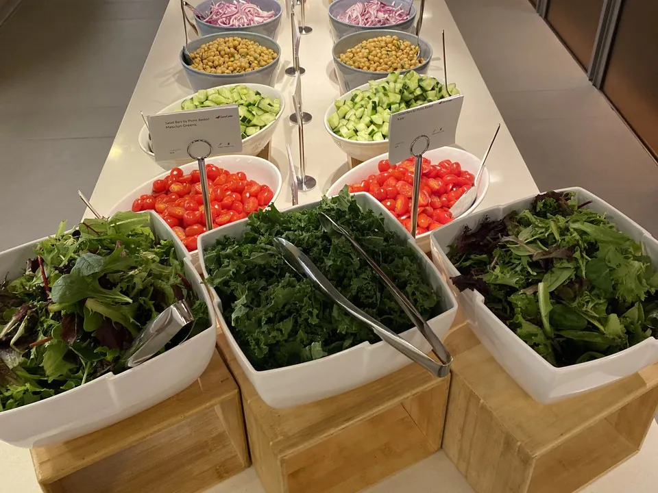 Fresh salad bar with bowls of mixed greens, kale, cherry tomatoes, cucumbers, chickpeas, and red onion on wood risers in an office
