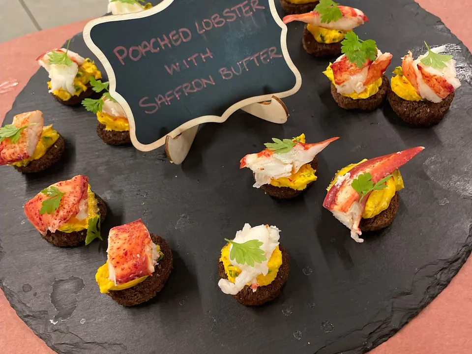 Poached lobster canapes on dark pumpernickel rounds topped with saffron butter and a microgreen, arranged on a black slate platter