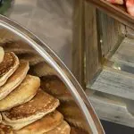 Stainless tray of golden buttermilk pancakes beside a wooden board of fresh strawberries, banana slices and blueberries on an office breakfast counter