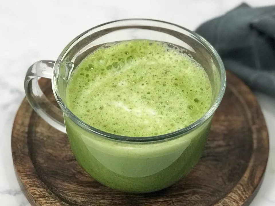 A frothy green matcha latte in a clear glass mug on a round wooden board with a gray linen napkin