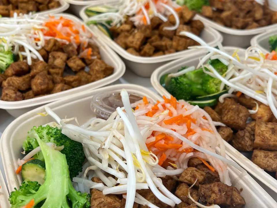Rows of individually packaged boxed meals with tofu, broccoli, bean sprouts, and pickled vegetables lined up for a conference-room working lunch