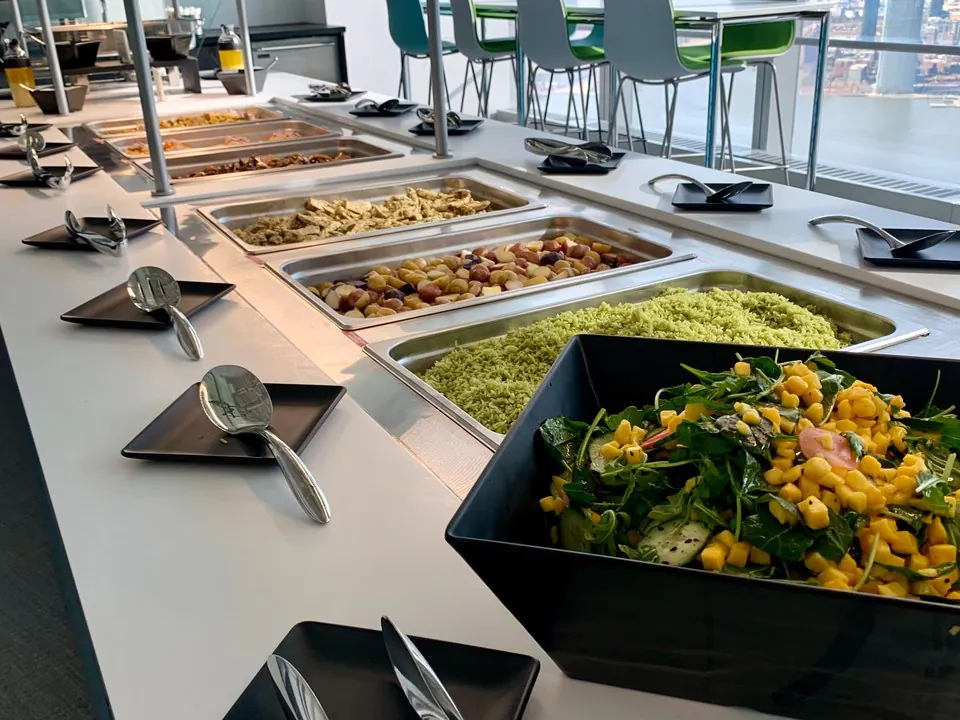 Office buffet line with chafing pans of pasta, potatoes, and chicken plus a large mixed green salad and black serving plates, the served-buffet option for a larger or more casual lunch and learn