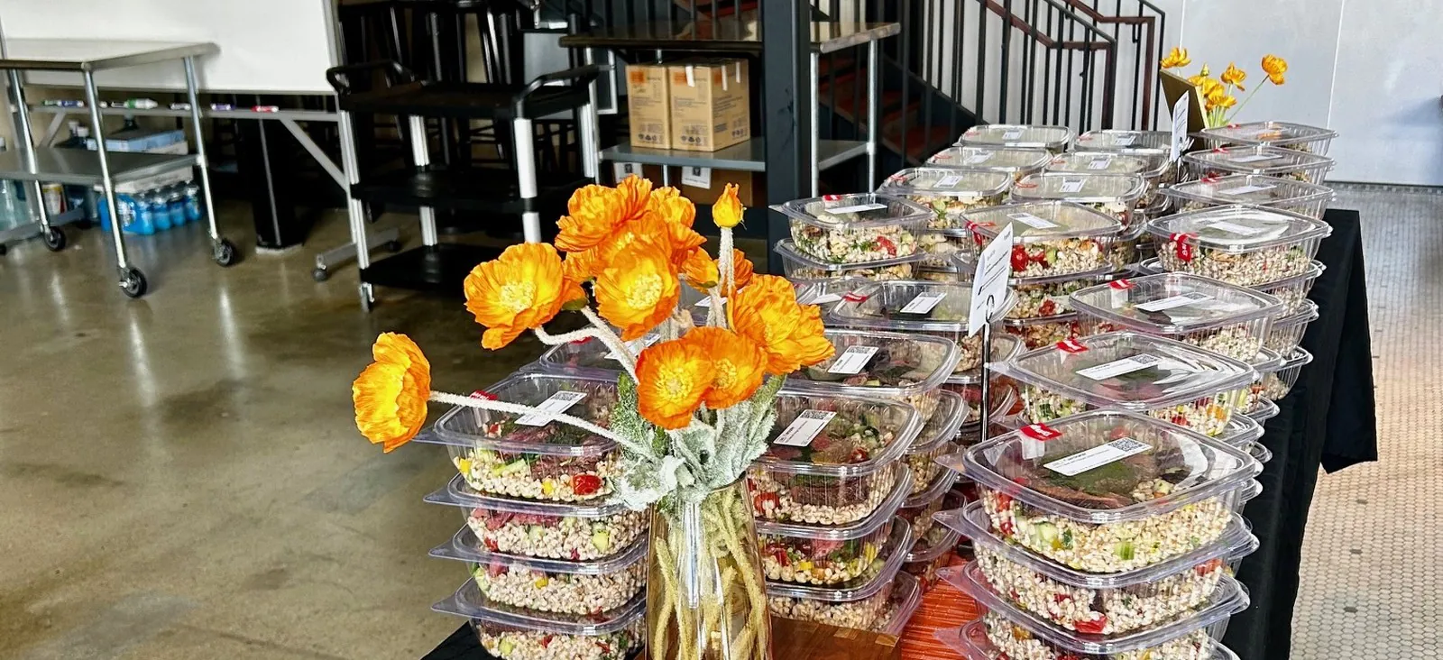 Rows of individually boxed grain salads staged on a meeting-room table with an orange floral centerpiece, the no-mess eat-and-listen format scaled for a lunch and learn session