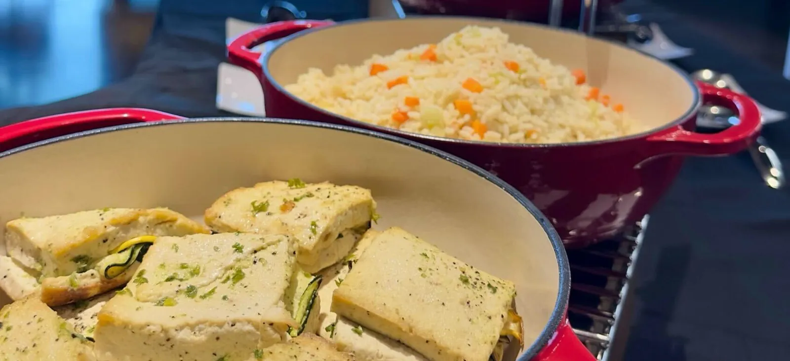 Red enameled dutch ovens of herbed chicken and rice pilaf with vegetables on an office buffet rack, a hearty hot homestyle catering spread