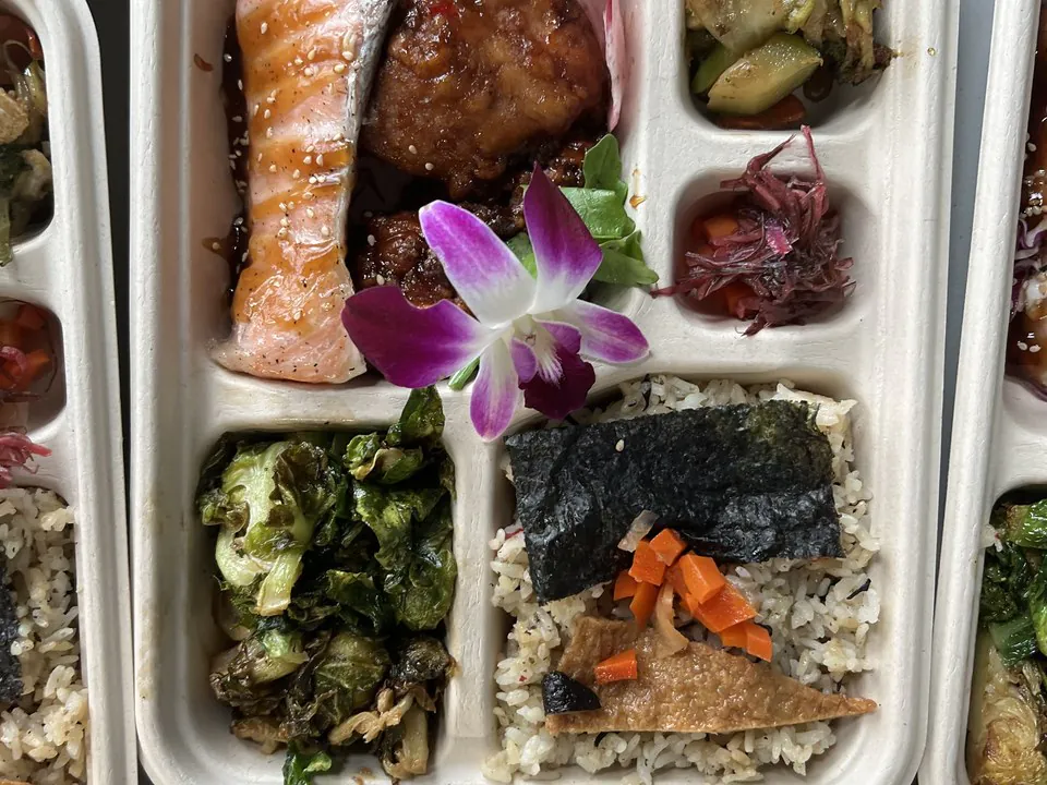 Overhead view of bento boxes with glazed salmon, teriyaki chicken, roasted brussels sprouts, and nori-topped rice