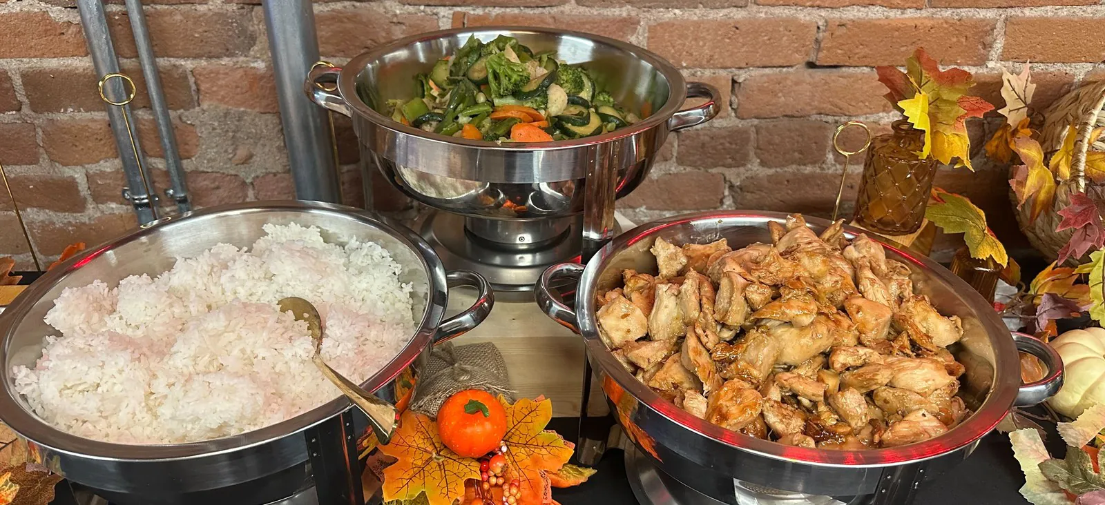 Office Asian catering buffet with chafing bowls of steamed rice, sauteed vegetables, and grilled chicken against a brick wall