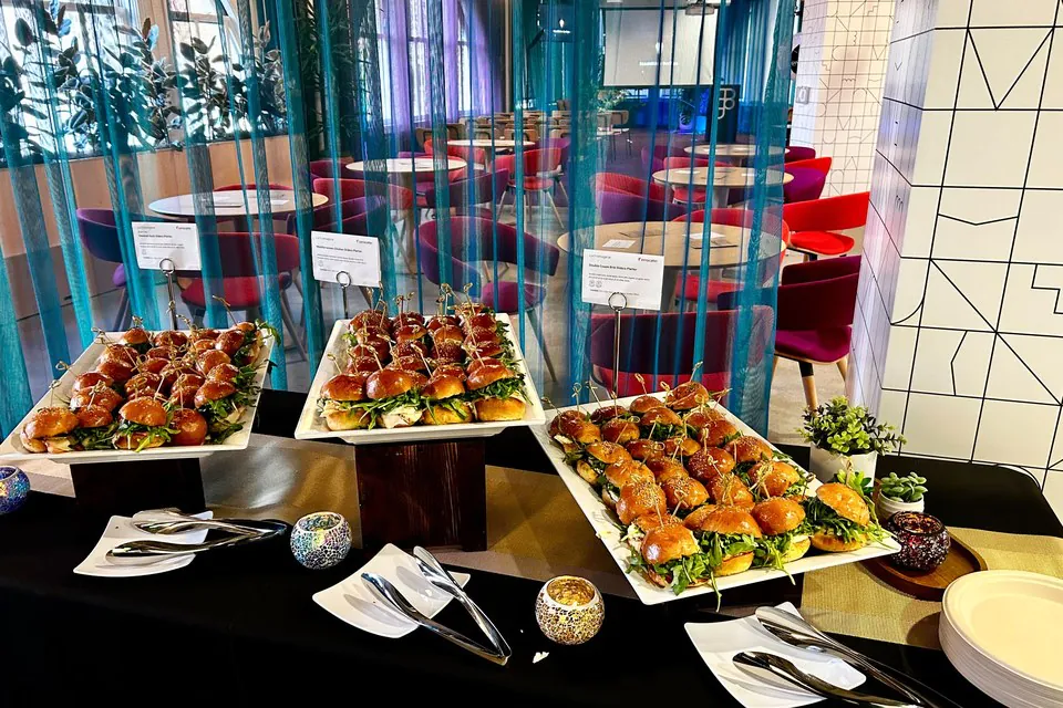 Three white catering platters of mini brioche slider sandwiches with toothpick picks on a black linen office table