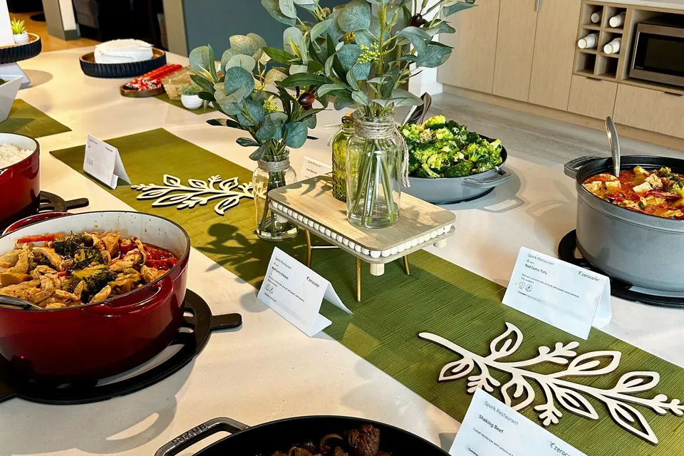 A styled office pasta spread with a red enameled pot of red-sauce pasta, a green salad bowl, and a eucalyptus table runner