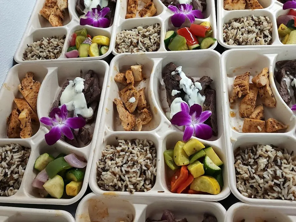 Grid of individual compartment meal boxes with grilled chicken, sliced steak, wild rice, and roasted vegetables, each topped with an orchid