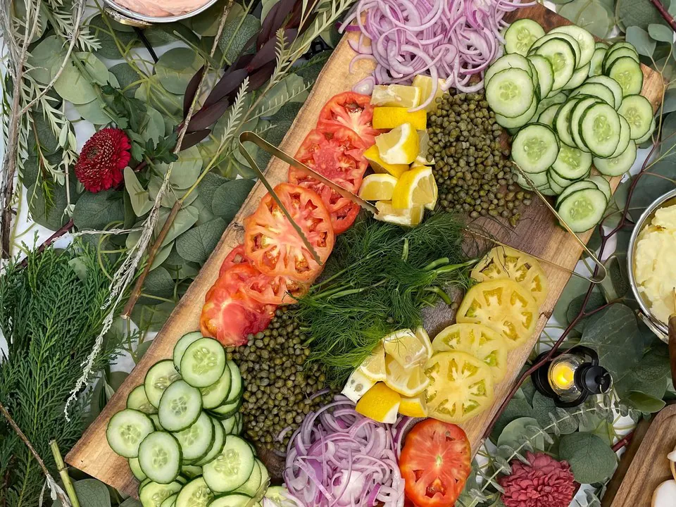 Build-your-own sandwich board with fresh sliced heirloom tomatoes, cucumbers, red onion, capers, lemon, and spreads on a wooden board