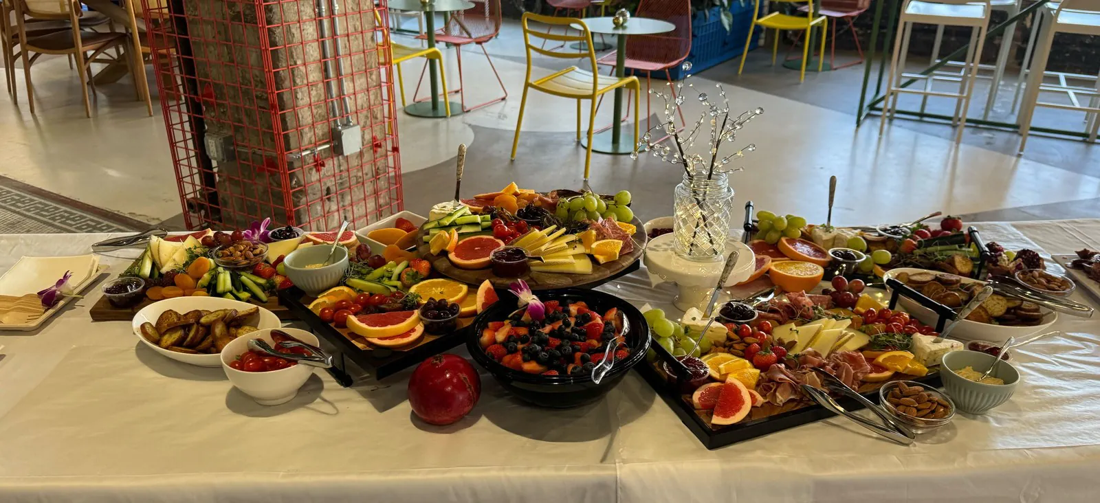 Abundant office catering spread of fresh fruit, charcuterie, and cheese platters on a long table in a bright office, with a red column accent