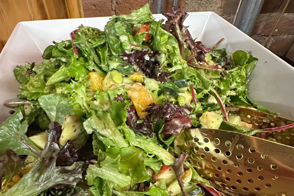 A close-up of a freshly tossed mixed-greens salad with mandarin oranges and edamame in a white square bowl, the kind of bowl a guest builds at a salad bar