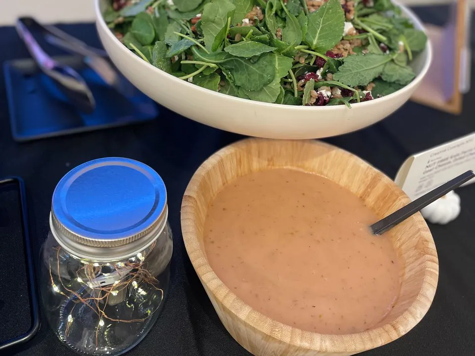 A bowl of kale and arugula salad with cranberries and feta set next to a separate wooden bowl of dressing with a ladle, on a build-your-own salad bar