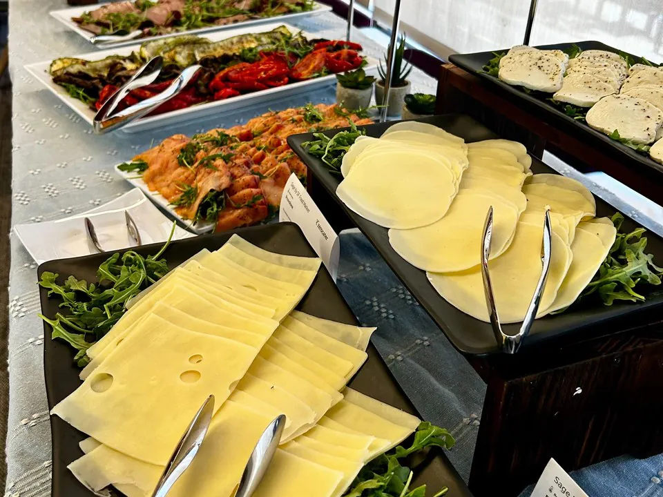 Build-your-own sandwich bar proteins and cheeses station with platters of sliced swiss, provolone, and mozzarella, grilled vegetables, and sliced meats on arugula with serving tongs