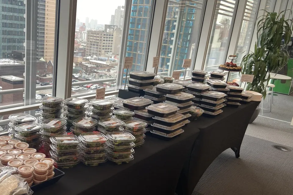 Stacks of individual boxed lunches and sauce cups on a draped table by a high-rise window for Seattle office catering