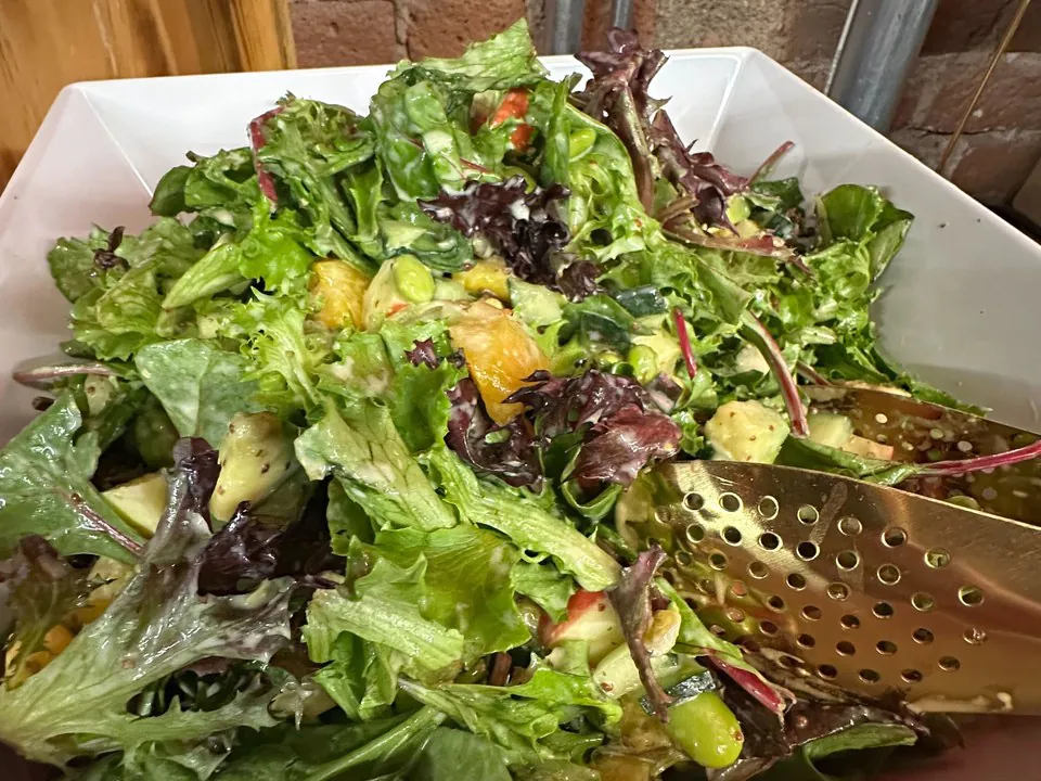 Close-up of a fresh mixed-greens salad with mandarin oranges, edamame, and apple in a white bowl with serving tongs