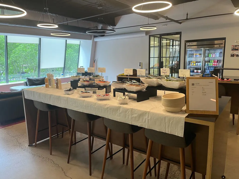 Build-your-own bowl bar on a long communal office table with bar stools, topping bowls, and a drinks fridge in the background