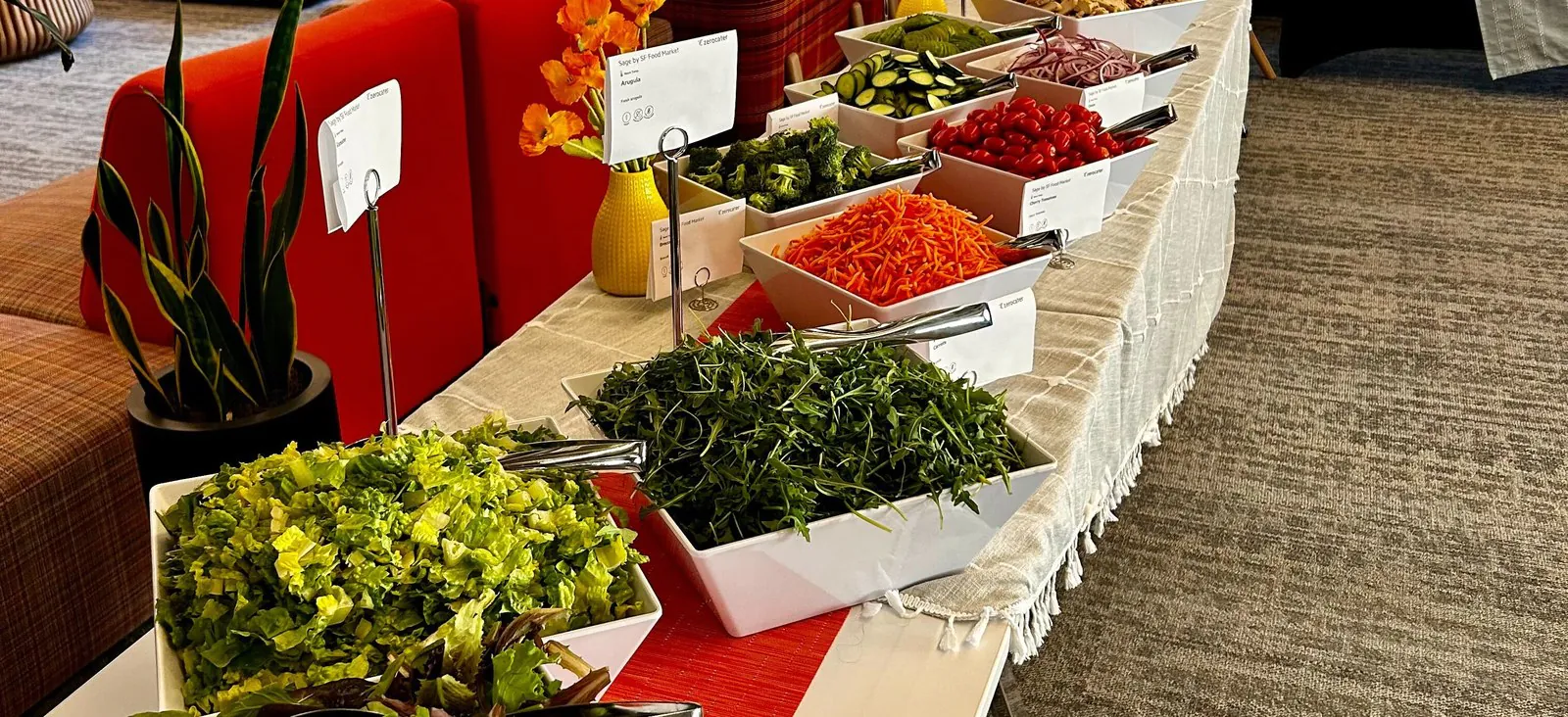 Abundant office salad bar with white bowls of mixed greens, kale, shredded carrots, peppers, and cucumbers along a table with a red runner