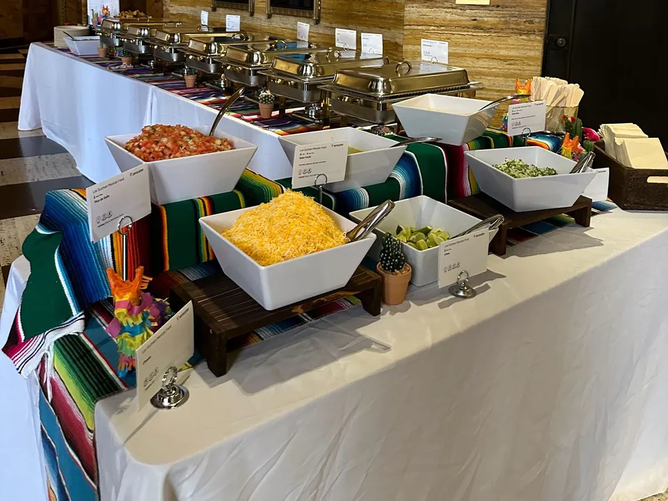 Build-your-own taco bar topping station with white bowls of red salsa, shredded cheese, diced jalapenos, and guacamole set on serape runners in front of chafing dishes