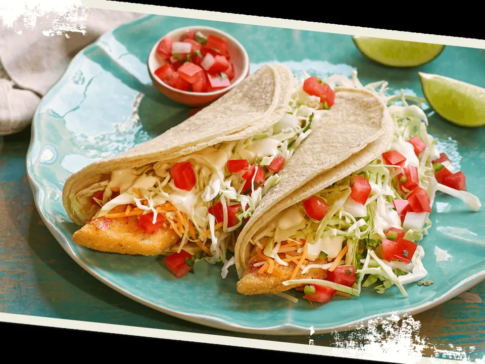Two Baja fish tacos with white fish, shredded cabbage, pico de gallo, and a bright crema sauce on a teal coastal plate with lime wedges