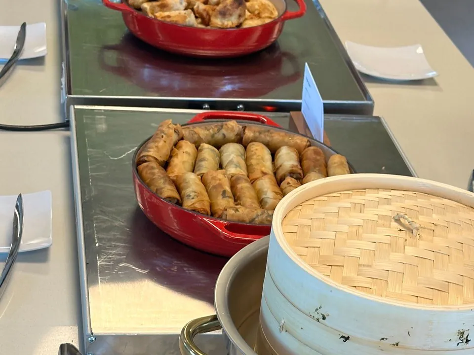 An Asian catering spread with red dishes of egg rolls and spring rolls, a chafing tray of fried rice, and bamboo steamers laid out on a buffet line