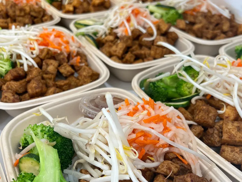 Rows of individual single-serve bowls with grilled tofu, fresh bean sprouts, shredded carrot, broccoli, and a cup of sauce