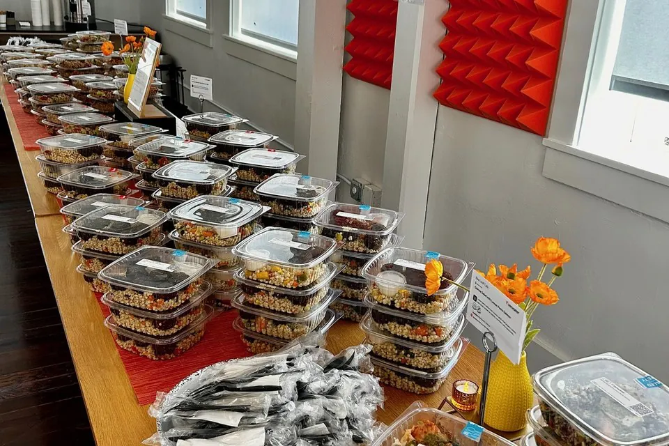 Rows of clear clamshell containers of fresh grain salads with wrapped utensil sets and orange flowers along a long catering table by a window