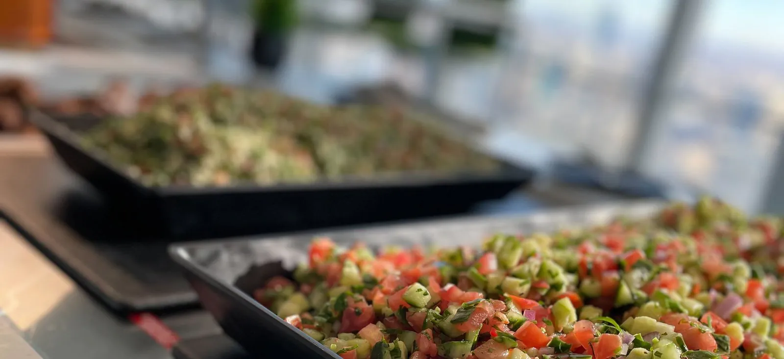 Office catering buffet with black trays of fresh chopped Israeli salad and tabbouleh on a counter beside a floor-to-ceiling window with a soft-focus city skyline behind