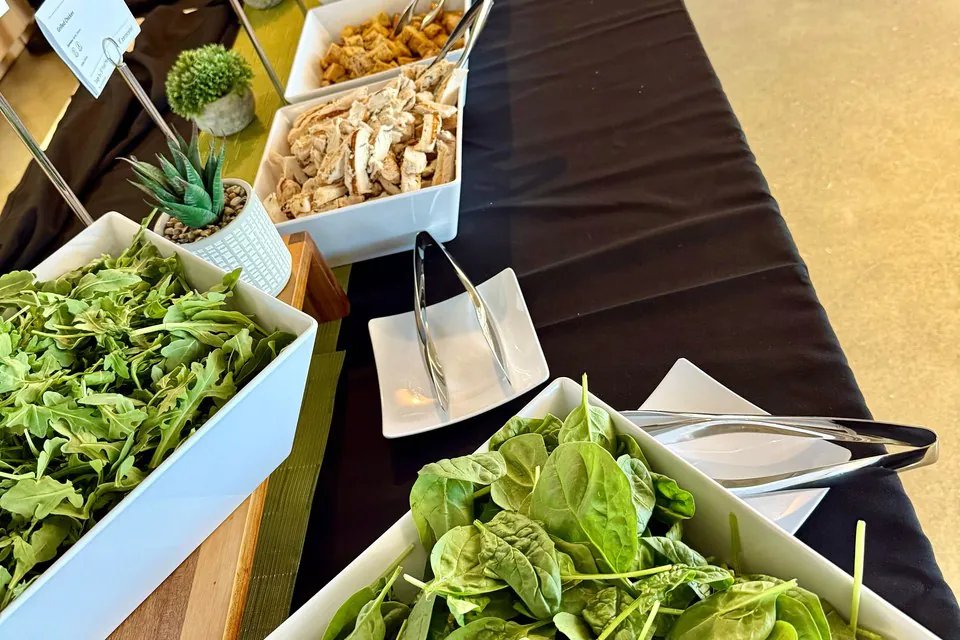 Build-your-own office salad bar with white square bowls of fresh spinach, arugula, sliced chicken, and toppings along a black-draped table with serving tongs