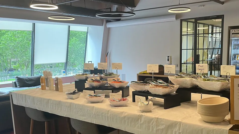 An office build-your-own bowl bar with topping trays, bowls of greens and vegetables, stacked serving bowls, and menu cards on a linen-draped table by floor-to-ceiling windows