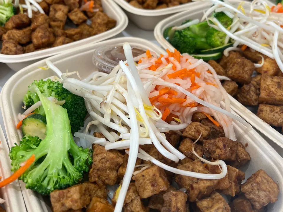 Close-up of plant-based bowls with tofu, bean sprouts, broccoli, and shredded carrot over rice in compostable containers