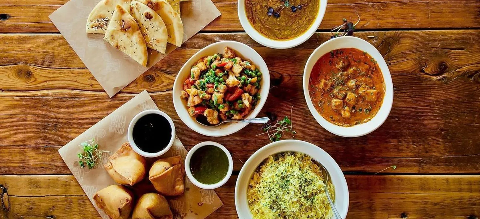 Curry Up Now office catering spread Curry Up Now Indian street food catering spread with samosas, chutneys, naan, paneer tikka masala, dal, and fenugreek rice on a wood table