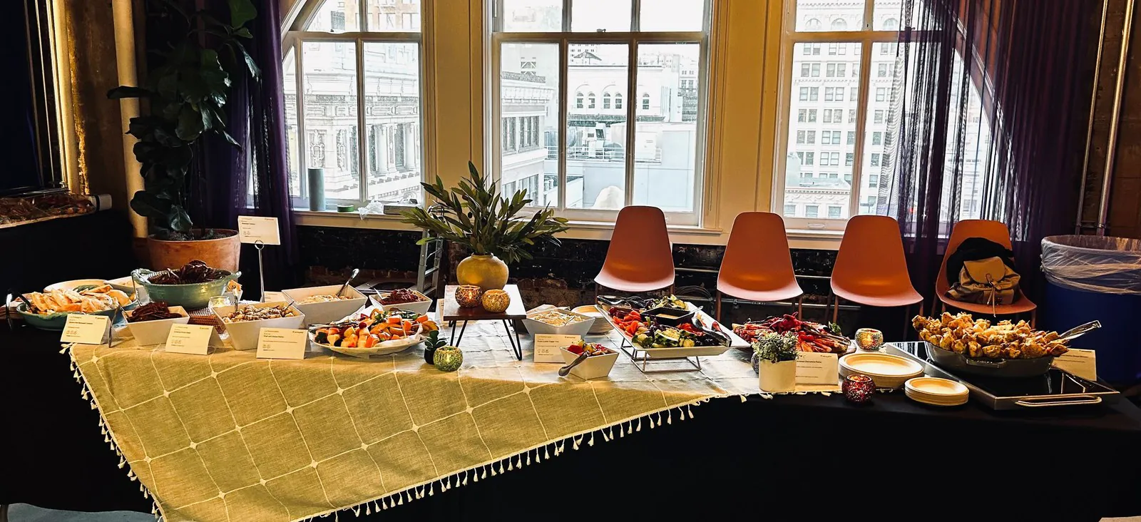 Catered buffet spread of salads, fruit, and hot appetizers on a long table in a downtown high-rise office with arched windows