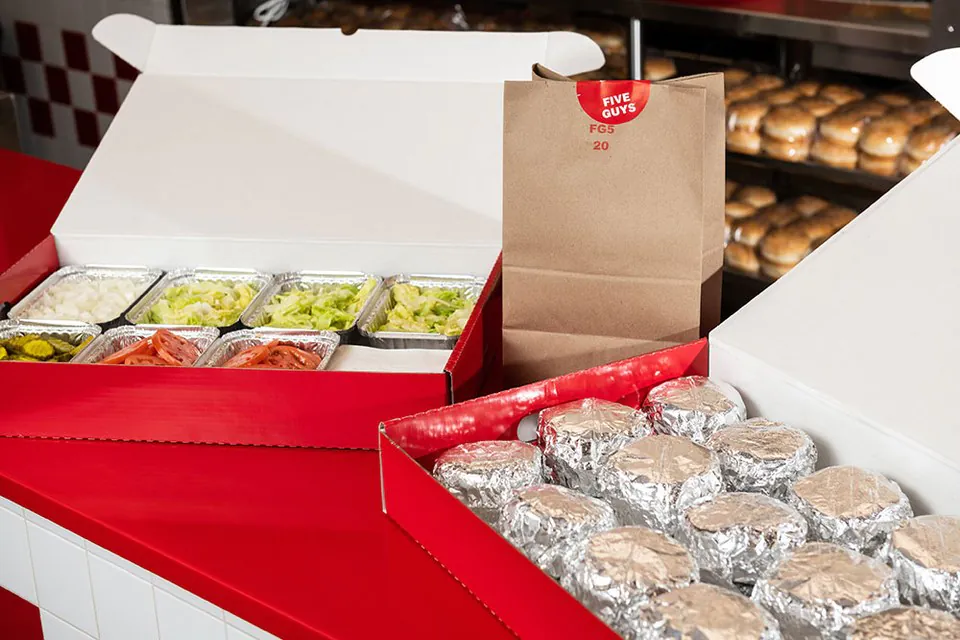 Five Guys catering boxes on a red counter: foil-wrapped burgers in one tray, onions, lettuce, pickles and sliced tomatoes in separate foil trays, with a branded paper bag and racks of buns behind