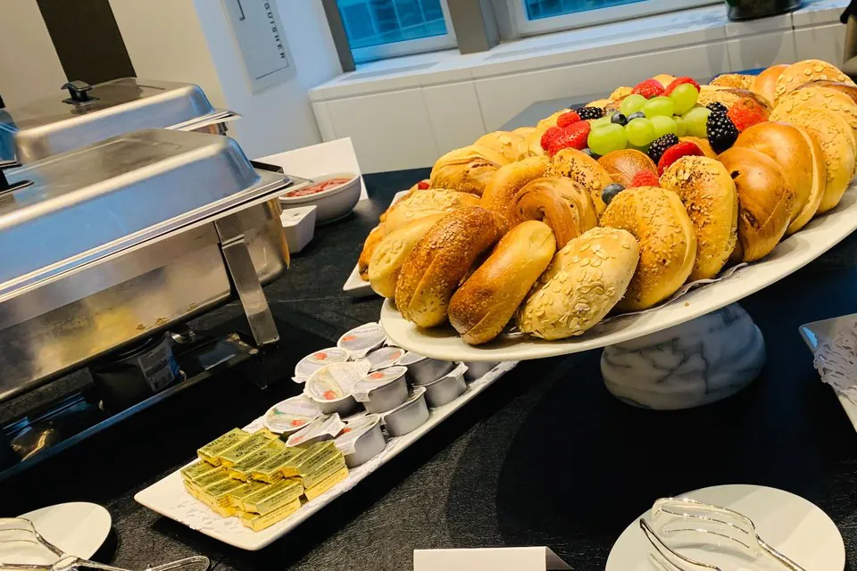 Office breakfast bagel platter topped with fresh fruit on a marble pedestal beside jam jars, butter, and a chafing dish