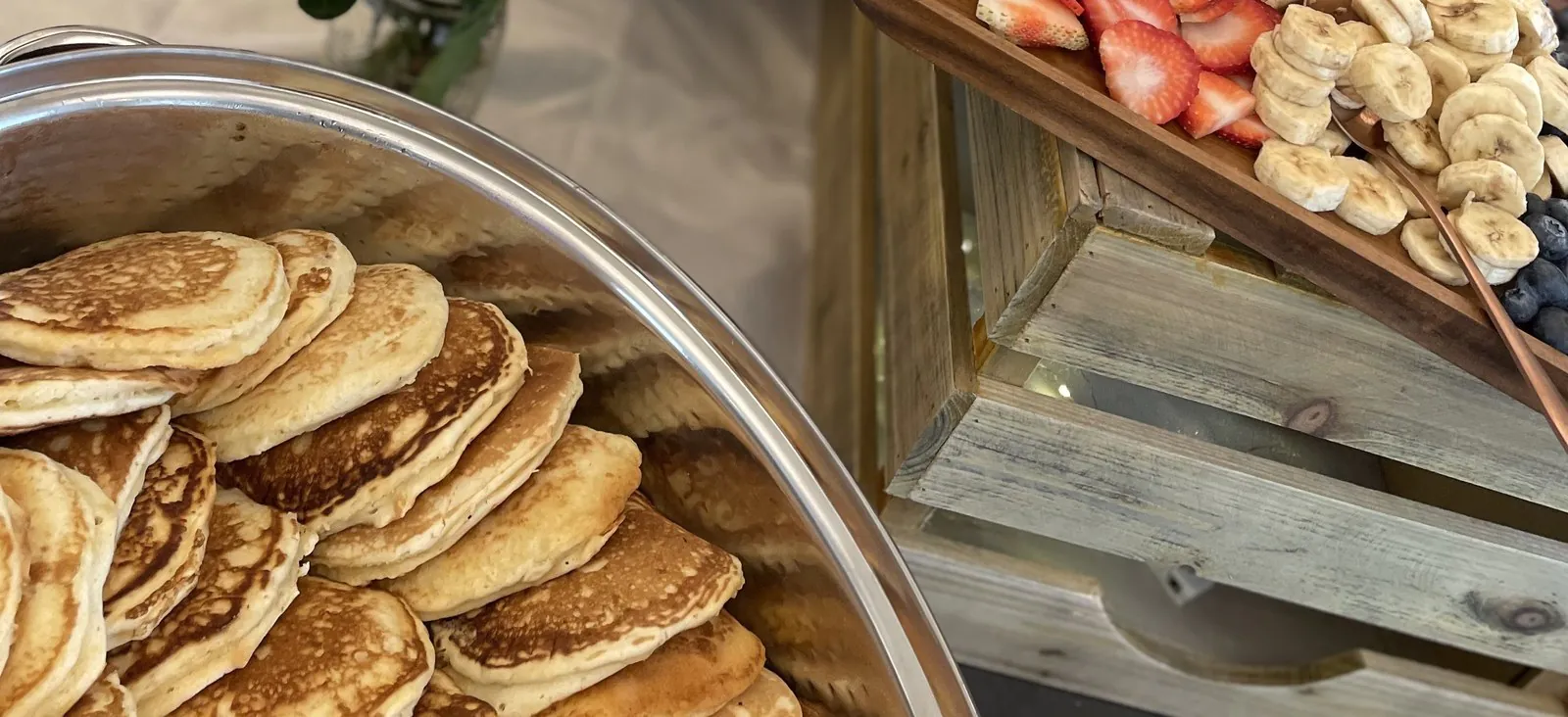 Office breakfast catering spread with a silver tray of golden griddle pancakes beside a wooden board of sliced strawberries, bananas, and blueberries
