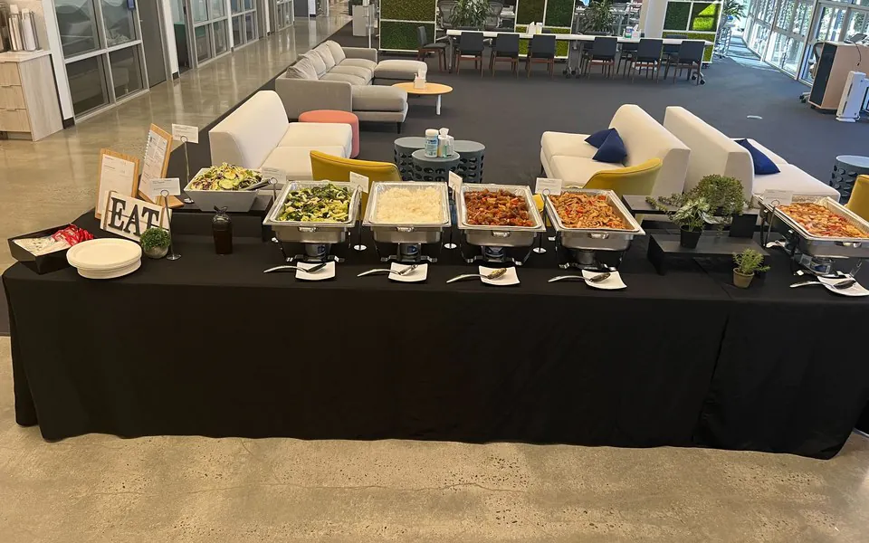 Hot chafing-tray buffet line of greens, rice, and saucy proteins set up on a long table for a plant crew