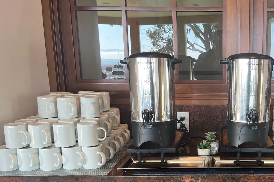 Offsite coffee break station with two stainless coffee urns and stacked white mugs on a counter overlooking the water