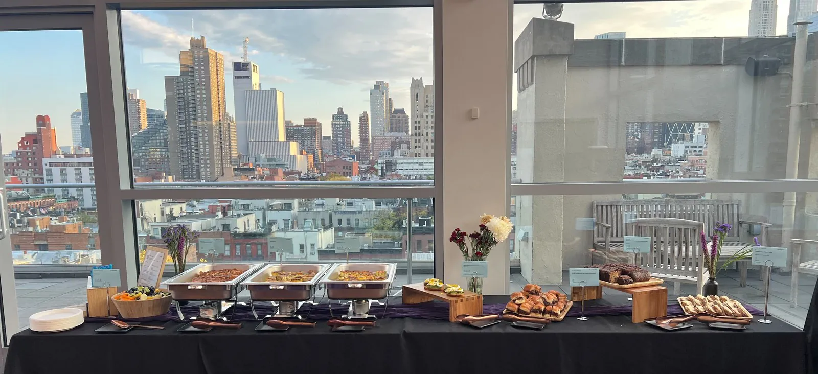 Offsite buffet line with chafing dishes, pastries and fruit on a black-draped table in an event space with a city skyline through the windows